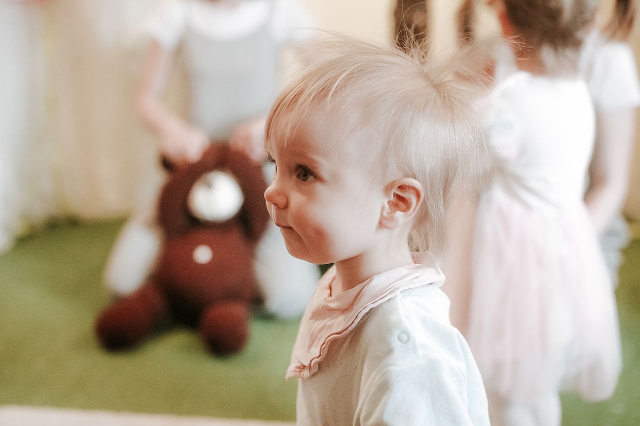 A cute toddler with blonde hair stands indoors, surrounded by toys and friends.